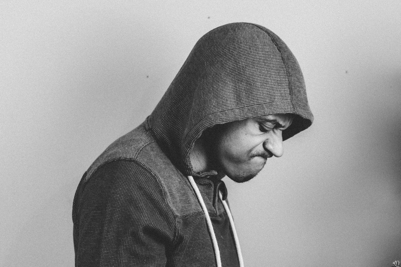 Siyah beyaz kapüşonlu bir ceket giyen bir adamın yakın çekimi. Kameradan hafifçe uzağa bakmaktadır ve yüzü ceketinin kapüşonu tarafından kısmen gizlenmiştir. Sağ eli çenesine yakın, sol eli ise göğsünün yanında durmaktadır. Kapüşonu yüzünün üst kısmına ve alnına gölge düşürüyor. Oldukça uzun boylu ve ince yapılı görünmektedir. Ceketin kapüşonu hafifçe kaldırılmış ve kapüşonun kenarları yukarı doğru çevrilmiş. Kapüşonlu ceketin kumaşının dokusu ve deseni net bir şekilde görülebiliyor.