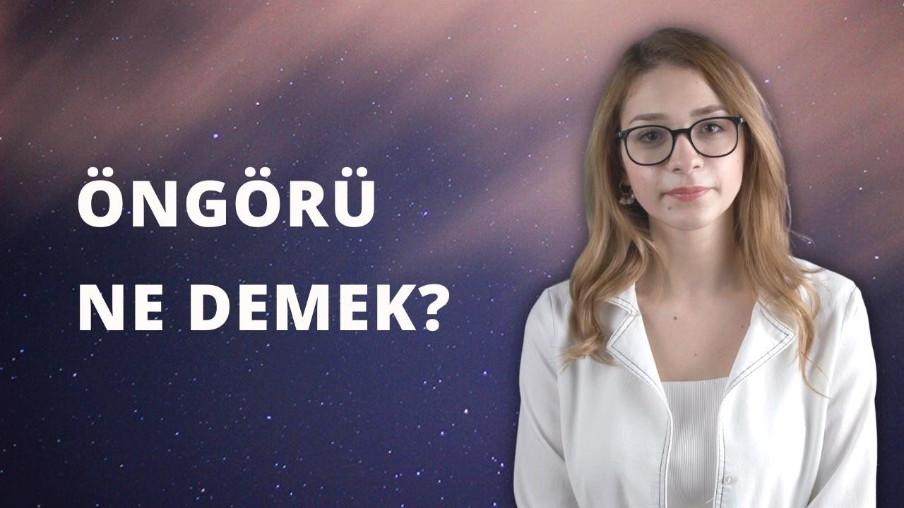 Gözlüklü ve beyaz gömlekli bir kadın doğrudan kameraya bakar. Gözlerinin çoğunu kapatan büyük, yuvarlak gözlükleri var. Dudakları hafifçe bükülmüş, dişleri zar zor görünüyor. Yüzünün sağ tarafında küçük bir güzellik izi vardır ve saçları yüzünden geriye doğru toplanmıştır. Beyaz gömleğinin yakası v yaka ve omuzlarından gevşekçe sarkıyor. Arka plan nötr bir renk ve çerçevede başka bir şey yok. Kadın kendinden emin ve kontrollü görünüyor.