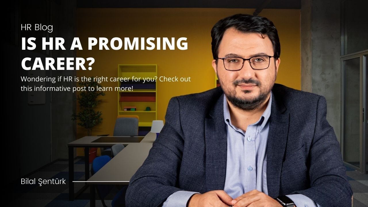 A man wearing a smart black suit stands in front of a yellow shelf. He has glasses on and a well-trimmed beard. His shirt is buttoned up and his eyes are visible in a close-up shot. He is looking ahead, with a blurry lamp in the background. To his right, there is a black and white sign with white lettering. On the shelf is an array of colorful objects.