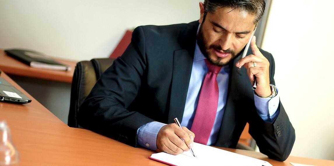 İş kıyafeti giymiş ve kravat takmış bir adam telefonla konuşuyor. Odaklanmış ve profesyonel görünüyor. Kravatı açık pembe renktedir ve takım elbisesinin kumaşı siyahtır. Yakındaki bir cam yüzeyde ışık yansıması görülüyor. Arka planda bir dizüstü bilgisayar görülüyor ve bir el kalem tutmuş, bir kağıda yazı yazarken görülüyor. Ön planda beyaz bir yüzey, arka planda bir telefonun yakın çekimi, bulanık. Takım elbiseli adam derin bir konsantrasyon içinde telefonla konuşuyor gibi görünüyor.