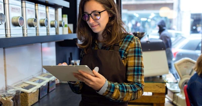 Kahverengi önlüklü bir kadın elinde bir tablet tutuyor. Yüzünde gözlük var ve dikkatle cihaza bakıyor. Ağzı hafifçe açık ve gözleri ekrana odaklanmış durumda. Sol eli cihazın arkasında dururken tableti sağ elinde tutuyor. Arka plan bulanık ve bir sıra metal kutu içeriyor. Kadının omzu, sağ alt köşede yüzünün yakın çekimiyle birlikte görüntüde görülebiliyor. Tableti tutarken kolu hafifçe bükülmüş ve sıcak bir gülümseme takınmış.