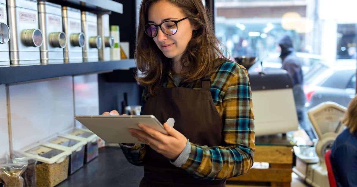 Kahverengi önlüklü bir kadın elinde bir tablet tutuyor. Yüzünde gözlük var ve dikkatle cihaza bakıyor. Ağzı hafifçe açık ve gözleri ekrana odaklanmış durumda. Sol eli cihazın arkasında dururken tableti sağ elinde tutuyor. Arka plan bulanık ve bir sıra metal kutu içeriyor. Kadının omzu, sağ alt köşede yüzünün yakın çekimiyle birlikte görüntüde görülebiliyor. Tableti tutarken kolu hafifçe bükülmüş ve sıcak bir gülümseme takınmış.