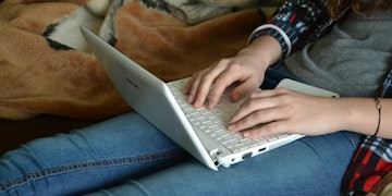 Bir kişi masa üzerinde dizüstü bilgisayar kullanıyor. Elleri klavyenin üzerinde ve gözleri ekrana odaklanmış durumda. Dizüstü bilgisayar gümüş rengindedir ve kişinin elleri klavye üzerinde görünmektedir. Tuşlar beyazdır ve dizüstü bilgisayar ekranı aydınlatılmıştır. Kişi yazı yazıyor ve parmakları hızla hareket ediyor. Dizüstü bilgisayarda bilgi içeren bir web sayfası ve sol üst köşede bir arama çubuğu görüntülenmektedir. Kişi kendini tamamen işine kaptırmış gibi görünüyor. Masa açık ahşap rengindedir ve kişi mavi bir gömlek giymektedir. Arka plan bulanık ve odak dışıdır.