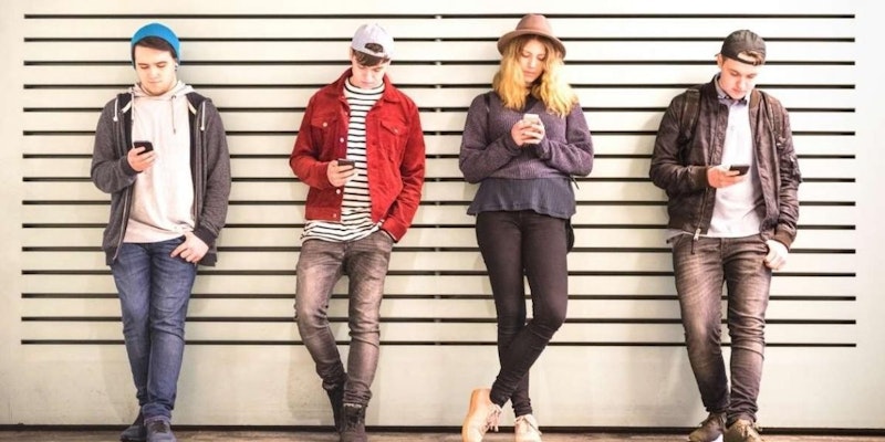 Bir duvarın önünde duran bir grup insan. Kırmızı ceketli bir adam, elinde bir telefon tutmaktadır. Adamın sağında şapkalı ve uzun saçları görünen bir kadın duruyor. Resmin sol tarafında elinde telefon tutan bir adam ve onun yanında duran bir kadın. Ortada bir kişinin başı hafifçe bulanıklaştırılmış. Tüm insanların duvarın önünde birlikte durdukları görülüyor.