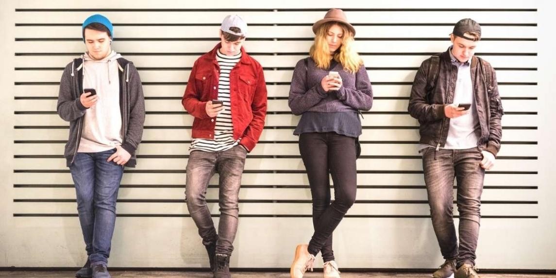 Bir duvarın önünde duran bir grup insan. Kırmızı ceketli bir adam, elinde bir telefon tutmaktadır. Adamın sağında şapkalı ve uzun saçları görünen bir kadın duruyor. Resmin sol tarafında elinde telefon tutan bir adam ve onun yanında duran bir kadın. Ortada bir kişinin başı hafifçe bulanıklaştırılmış. Tüm insanların duvarın önünde birlikte durdukları görülüyor.