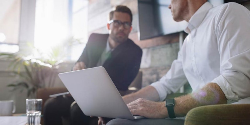 Takım elbiseli ve kravatlı bir adam yuvarlak bir masa üzerinde dizüstü bilgisayar kullanıyor. Yüzü yakın plandadır ve dikkatle dizüstü bilgisayarın ekranına bakmaktadır. Dizüstü bilgisayarın etrafı bulanık beyaz bir kupa ve seçilmesi zor yeşil bir nesneyle çevrili. Gıcır gıcır beyaz bir gömlek giyen adamın yüz hatları keskin ve nettir. Odaklanmış ve kararlı görünüyor.