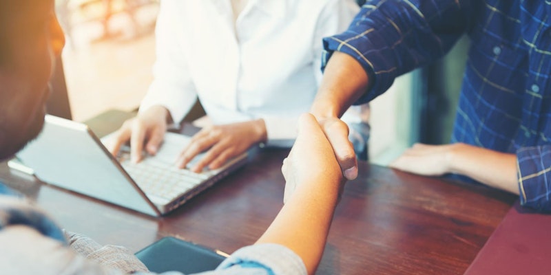 İki kişi yakın çekimde, elleri tokalaşmak üzere kenetlenmiş olarak görülüyor. Her iki kişi de resmi kıyafetler giymiş ve yüzlerinde ciddi bir ifade var. Soldaki adam lacivert bir takım elbise giymiş ve açık mavi bir kravat takmıştır. Kısa siyah saçları ve keskin bir çene hattı vardır. Sağdaki kadın krem rengi bir bluz ve açık gri bir etek giyiyor. Saçlarını düzgün bir topuz yapmış ve bir çift küçük gümüş küpe takmış. Ellerini sıkıca birbirine bastırarak tokalaşıyorlar, bu bir saygı ve hayranlık jesti. Arka plan çoğunlukla belirsiz, ancak batan güneşin ışığı arka planda görülebiliyor ve sahneye yumuşak, sıcak bir ton veriyor.