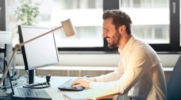 Beyaz gömlekli bir adamın yakın plan görüntüsü bilgisayar başında gülümsemektedir. Elleri klavyenin üzerindedir ve yakın çekimde sağ eli tuşların üzerindedir. Arka planda beyaz bir tahtanın yakın çekimi de görülüyor. Adam sakallıdır ve gözlük takmaktadır. Görüntünün sağ alt köşesinde bir bilgisayar ekranının yakın çekimi görülüyor ve klavye üzerinde bir kişinin elinin bulanık görüntüsü de görülebiliyor. Ayrıca, arka planda siyah bir duvarın yakın çekimi görülmektedir. Tüm bu unsurlar bir araya gelerek mutlu bir şekilde bilgisayar kullanan bir adamın detaylı görüntüsünü oluşturuyor.