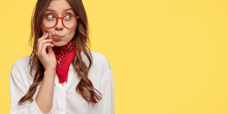 Gözlüklü ve kırmızı bandanalı bir kadın görülüyor. Yüzünde nötr bir ifade var ve doğrudan kameraya bakıyor. Saçları topuz yapılmış ve bandanası başının üst kısmını örtüyor. Bandana parlak kırmızıdır ve üzerinde beyaz bir desen vardır. Gözlükleri ince siyah bir çerçeveye sahip ve köşeleri hafif sivri. Beyaz bir tişört giymiş ve kollarını önünde kavuşturmuş. Arka plan bulanık, ancak uzakta yeşil bir manzaraya dair bir ipucu var. Sakin ve kendinden emin görünüyor ve gözleri uzaktaki bir şeye odaklanmış gibi görünüyor.