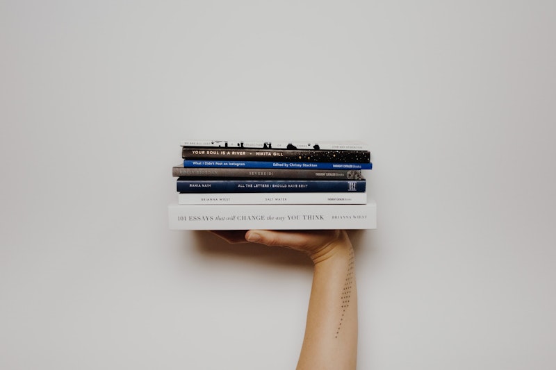 Bir kişi sol eliyle bir yığın kitap tutarken görülüyor. Kitaplar büyük ve kalındır, her birinin kapağında bir metin yazılıdır. Kişinin kolu görünüyor ve derisinde bir dövme görülebiliyor. Arka plan beyazdır ve kitapların üzerinde birkaç harfin görülebildiği siyah yazılar vardır. Kişi ayakta durmakta, vücudu hafifçe sola doğru eğilmiş ve kitap yığınına bakmaktadır. Parmakları kitapların etrafında kıvrılmış ve yığındaki kitaplar eşit aralıklarla dizilmiş.