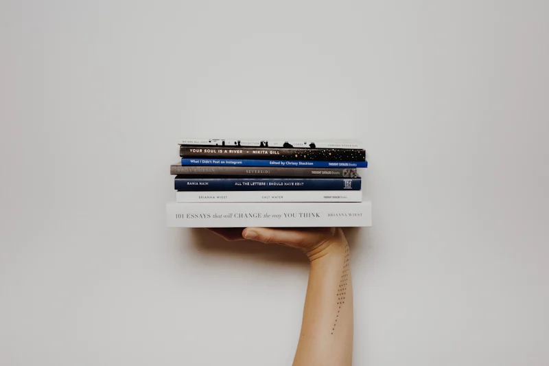 Bir kişi sol eliyle bir yığın kitap tutarken görülüyor. Kitaplar büyük ve kalındır, her birinin kapağında bir metin yazılıdır. Kişinin kolu görünüyor ve derisinde bir dövme görülebiliyor. Arka plan beyazdır ve kitapların üzerinde birkaç harfin görülebildiği siyah yazılar vardır. Kişi ayakta durmakta, vücudu hafifçe sola doğru eğilmiş ve kitap yığınına bakmaktadır. Parmakları kitapların etrafında kıvrılmış ve yığındaki kitaplar eşit aralıklarla dizilmiş.