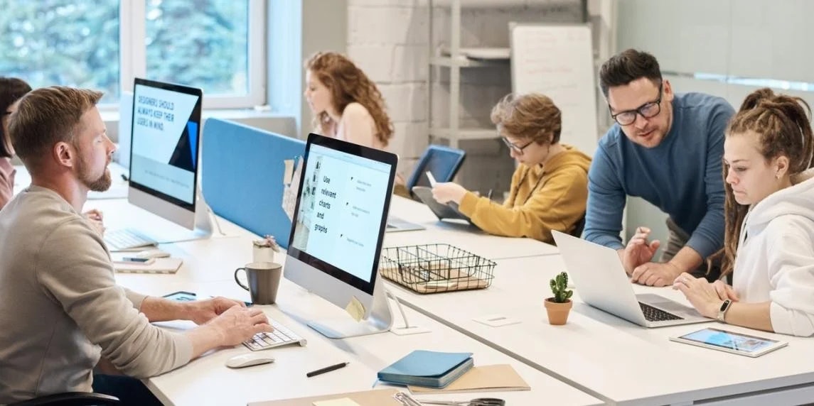 Bir grup insan bilgisayarlarla dolu bir odada bir masanın etrafında toplanmıştır. Ön tarafta gözlüklü ve mavi gömlekli bir adam oturmakta, saçları rastalı bir kadın ise bir kâğıda bakmaktadır. Masanın ortasında, içinde bir kağıt parçası bulunan tel bir sepet var. Masanın sol tarafında, bilgisayar ekranında yazılar görünen bir dizüstü bilgisayar var. Sağ tarafta ise içinde bilgisayar olan mavi bir çanta var. Herkes yoğun bir şekilde bilgisayarda çalışıyor, bazıları yazı yazıyor, bazıları da kitap okuyor.