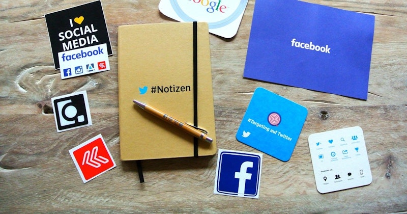 Bu görselde bir masanın üzerinde kalem, çıkartmalar ve diğer logoların bulunduğu bir defter yer almaktadır. Defter açık ve ortasında beyaz bir 'f' logosu olan mavi bir kare görülüyor. Defterin solunda üzerinde oklar bulunan kırmızı ve beyaz bir çıkartma görülüyor. Defterin sağında beyaz metinli ve birkaç logolu siyah bir işaret vardır. Ön planda, kapağı çıkarılmış bir kalemin yakın çekimi var. Resmin sağ üst köşesinde beyaz metinli mavi bir kağıt var. Sağ alt köşede, üzerinde birkaç mavi simge bulunan kare şeklinde beyaz bir kağıt var. Bu görüntünün renkleri ve dokuları, mevcut nesnelerin canlı ve ayrıntılı bir görünümünü sağlamaktadır.