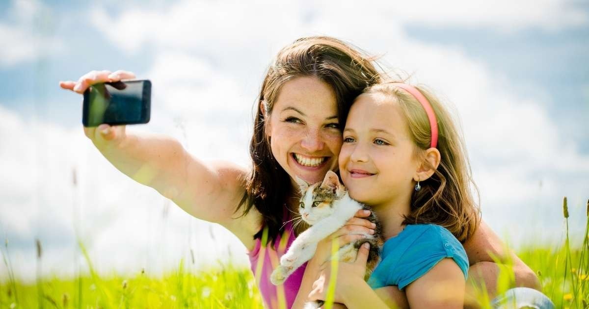 Bir kadın ve genç bir kız selfie çekerken, kız göğsüne yakın beyaz ve gri bir kedi tutuyor. Kadın mavi bir gömlek giyiyor ve saçında bir saç bandı var. Her ikisi de kameraya doğru gülümsüyor ve kız gözlük takıyor. İyi aydınlatılmış bir odadalar ve çerçeve parlak renklerle dolu. Kadın kolunu kıza dolamış ve doğrudan kameraya bakıyor. Kızın başı hafifçe yana eğiktir ve gülümserken gözleri kapalıdır.