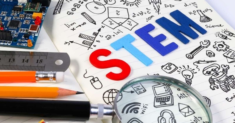 Kalemler ve bir büyüteçle birlikte bir defterin yakın çekimi görüntülenir. Defter açık ve sayfada çeşitli çizimlerin yanı sıra kırmızı bir harf ve üst köşede yazılı mavi ve kırmızı bir 'T' harfi var. Sayfanın üzerine bir büyüteç ve onun üzerine de bir cetvel yerleştirilmiştir. Arka planda siyah ve turuncu bir nesnenin yakın çekimi ve bir devre kartının yakın çekimi var. Her şey beyaz bir arka plana karşı, görüntüye net ve keskin bir odak sağlıyor.