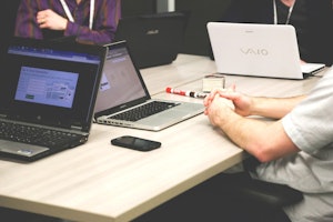 Bir adam iki dizüstü bilgisayar, siyah bir cep telefonu ve bir kalemin önünde bir masada oturuyor. Beyaz bir gömlek giymiş ve ellerini kucağında kavuşturmuş. Kendisine en yakın olan dizüstü bilgisayarın ekranında bir web sayfası açık. En uzağındaki dizüstü bilgisayarın üzerinde bir tablet vardır. Arka planda, elinde bir yığın kitap tutan bir kişinin bulanık bir görüntüsü var. Adamın oturduğu masa beyazdır ve dizüstü bilgisayarlar ile telefonu birbirine bağlayan çok sayıda kablo vardır.