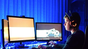Genç bir çocuk bilgisayarın önünde oturmakta ve kırmızı ışıklı bir çift kulaklık takmaktadır. Dikkatle ekrana odaklanmıştır ve elleri klavyede hızla yazmaktadır. Arka planda yüzü bulanıktır ve ellerinden birinin yakın çekimi görülebilmektedir. Bilgisayar monitöründe beyaz bir ekran ve ön planda bir cihaz vardır. Arka planda bulanık bir motosiklet görüntüsü de var. Çocuk odada yoğun bir odaklanma ve konsantrasyonla çevrelenmiştir.