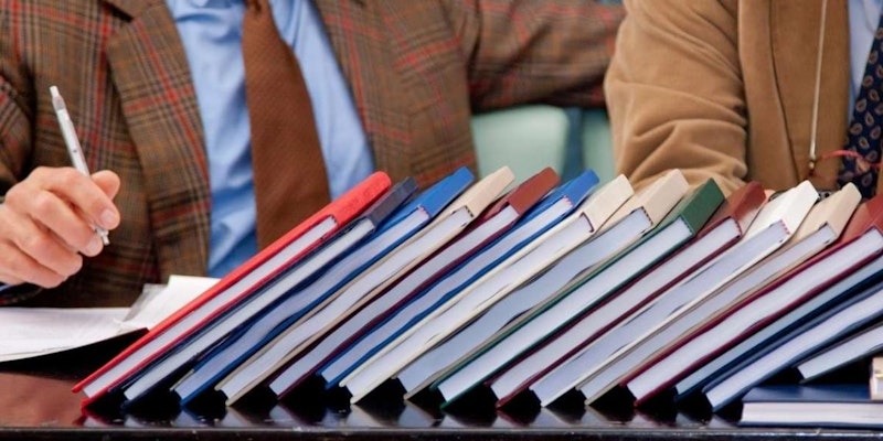 Resimde bir masanın üzerindeki kitap yığınının yakın çekimi gösterilmektedir. Yığın, çeşitli renk ve dokulara sahip çeşitli kitaplardan oluşmaktadır. Sağ alt köşede kalem tutan bir kişinin kolu görülüyor. Kitaplar düzgün, düzenli bir yığın halinde dizilmiş ve öndeki kitap diğerlerinden biraz uzağa doğru eğilmiş. Arka planda takım elbiseli ve kravatlı bir adam görülüyor. Kitapların ve adamın takım elbisesinin renkleri nötr arka planla kontrast oluşturuyor. Görüntü, kitapların ve adamın takım elbisesinin ayrıntılarını vurgulayan bir yakın çekimdir.