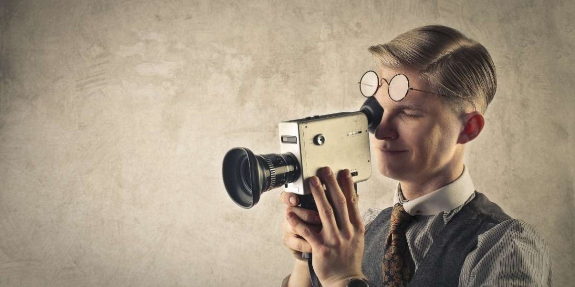 Bu resim elinde fotoğraf makinesi olan bir adamı gösteriyor. Arka planı beyaz olan iyi aydınlatılmış bir odada duruyor. Başı hafifçe yana eğik ve doğrudan kameraya bakıyor. Üzerinde kolları kıvrılmış beyaz bir gömlek ve siyah bir yelek vardır. Sol elinde, objektifi kameraya doğru bakan siyah bir kamera tutmaktadır. Sağ eli kameranın gövdesinin üzerindedir. Resmin sağ alt köşesinde bir ayakkabı var. Ayakkabı, yan tarafında bağcık deseni olan siyah bir elbise ayakkabısıdır.