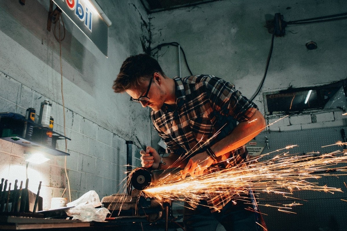 Koruyucu gözlük ve yüz maskesi takan bir adam metal bir nesneyi kesmek için açılı taşlama makinesi kullanıyor. Çeşitli sprey kutularının bulunduğu bir raf da dahil olmak üzere çeşitli alet ve malzemelerin bulunduğu bir tezgahın önünde duruyor. Görüntünün sağ üst köşesinde beyaz bir nesne görülüyor, ancak odak dışında ve seçilmesi zor. Adam işine odaklanmış, çalışırken kıvılcımlar uçuşuyor. Sahne öğütücünün parlak ışığıyla aydınlanıyor.