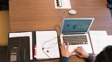 Siyah gömlek giyen bir kişi dizüstü bilgisayarının başında oturmuş, iki eliyle klavyede bir şeyler yazmaktadır. Dizüstü bilgisayar açık ve ekranı aydınlık. Dizüstü bilgisayarın sol tarafında, içinde kağıtlar ve bir çift kulaklık bulunan siyah bir klasör vardır. Sağ tarafta beyaz dikdörtgen bir nesne ve üzerinde birkaç çıkış bulunan dikdörtgen bir panel vardır. Dizüstü bilgisayarın solunda kare tasarımlı beyaz dairesel bir nesne vardır. Arka planda bir cep telefonu taslağına bakan bir kişi var. Son olarak, dizüstü bilgisayarın önündeki ahşap masanın üzerinde bir hesap makinesi ve bir klasör içinde kağıtlar var.