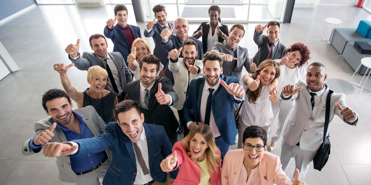 Bir arada duran ve mutlu bir şekilde gülümseyen bir grup insan. Grup çoğunlukla yetişkinlerden oluşuyor, aralarında çocuklar da var. Grubun ortasında açık mavi bir elbise giymiş ve parmağıyla yukarıyı işaret eden bir kadın var. Kadının sağında takım elbiseli bir adam başparmağıyla işaret ediyor ve gülümsüyor. Solunda pembe bir elbise giymiş ve selfie çeken başka bir kadın var. Takım elbiseli adamın sağında ise yine takım elbiseli ve mavi gömlekli bir başka adam başıyla onaylıyor. Ön planda bir grup erkek gülüyor ve kollarını uzatıyor. İçlerinden birinin kravat taktığı, görüntünün yakın plan çekiminde görülebiliyor. Arka plandaki insanlar bir arada duruyor, bazıları gülümsüyor ve diğerleri kameraya bakıyor.