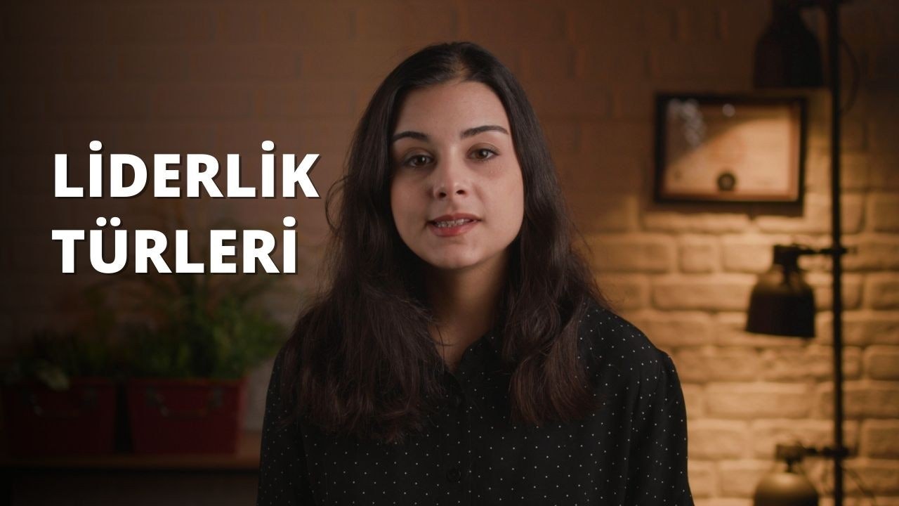 Siyah gömlek giyen uzun saçlı bir kadın kameranın önünde durmaktadır. Saçları geriye doğru toplanmış ve sırtından aşağıya doğru dökülüyor, uçları omuzlarının hemen üzerine kadar uzanıyor. Siyah gömleğinin kolları uzun ve düğmeleri boynuna kadar iliklenmiş. Yüzünde ciddi bir ifade var, gözleri doğrudan kameraya bakıyor. Arka planda bir bitkinin bulanık görüntüsü ve sağda beyaz bir metnin yakın çekimi var. Kadın tetikte duruyor, duruşu bir güven ve güç havası veriyor.