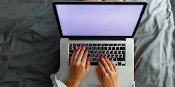 Bir dizüstü bilgisayarın önünde oturan bir kadının yakın plan görüntüsü. Mavi bir gömlek giymiş ve elleri klavyenin üzerinde. Gözleri ekrana doğru bakıyor ve muhtemelen konsantre olmuş bir şekilde ağzı hafifçe açık. Dizüstü bilgisayar açık ve klavyedeki tuşlar görülebiliyor. Dizüstü bilgisayar, üzerinde beyaz bir kupa ve birkaç başka eşya bulunan gri bir masanın üzerinde durmaktadır. Arka plan, köşesinde bir pencere olan düz beyaz bir duvardır. Kadın önemli bir şey üzerinde çalışıyor gibi görünüyor.