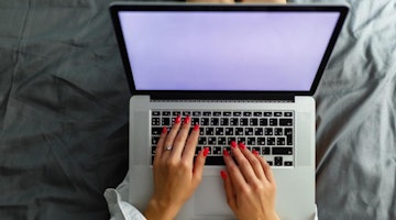 Bir dizüstü bilgisayarın önünde oturan bir kadının yakın plan görüntüsü. Mavi bir gömlek giymiş ve elleri klavyenin üzerinde. Gözleri ekrana doğru bakıyor ve muhtemelen konsantre olmuş bir şekilde ağzı hafifçe açık. Dizüstü bilgisayar açık ve klavyedeki tuşlar görülebiliyor. Dizüstü bilgisayar, üzerinde beyaz bir kupa ve birkaç başka eşya bulunan gri bir masanın üzerinde durmaktadır. Arka plan, köşesinde bir pencere olan düz beyaz bir duvardır. Kadın önemli bir şey üzerinde çalışıyor gibi görünüyor.