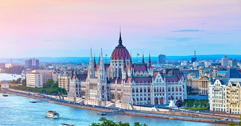 Görüntünün ön planında, yemyeşil ağaçlarla çevrili, kırmızı kubbeli büyük beyaz bir bina duruyor. Binanın çok sayıda penceresi ve kubbenin etrafında siyah bir süslemesi var. Arka planda, kenarına yakın bir yerde duran bir insanla birlikte geniş bir nehir görüntüyü boydan boya kat ediyor. Gökyüzü masmavi ve güneş sahnenin üzerinde parlıyor. Resmin tamamı çok huzurlu ve dingin, nehir ve bina güzel bir kontrast oluşturuyor.