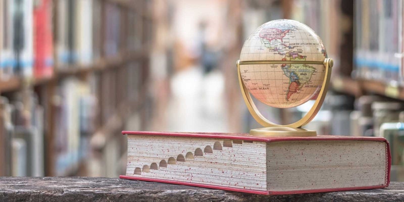 Bir küre, düz bir yüzey üzerinde duran bir kitabın üzerine yerleştirilmiştir. Küre, dünyanın kıtalarını tasvir eden, maviler ve yeşillerle dolu büyük, kalın bir haritadan oluşuyor. Altındaki kitap açık renkli bir ciltli, sırtı görünüyor ve ön yüzünden birkaç sayfa dışarı taşmış. Küre odak noktasındayken, kitap hafifçe odak dışında kalıyor ve yumuşak, bulanık bir görünüm veriyor. Görüntü, dünyanın ayrıntılarını ve kitabın üzerindeki yerleşimini sergileyen harika bir alan derinliğine sahip.