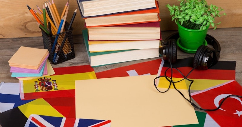 Ahşap bir masanın üzerinde bir yığın kitap ve bir bitki. Kitaplar parlak renkli ve düzgün bir yığın halinde dizilmiş, en üstteki görülebilmesi için hafifçe eğilmiş. Bitki yemyeşil ve aynı renkte bir saksıya sahip. Kitapların yanında bir kalemlik ve bir yığın renkli kâğıt var. Bitkinin yanındaki bayrağın üzerine bir çift siyah kulaklık yerleştirilmiş. Görüntü birkaç kitabın yakın çekimidir, arka planda yeşil saksı bitkisi ve kulaklık detayı vardır.