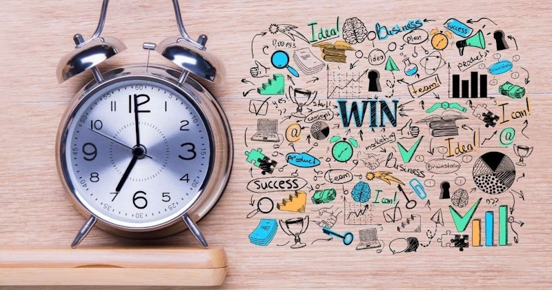 Ahşap bir yüzeyin üzerinde duran, siyah ve mavi romen rakamlı bir yüze sahip metal bir çalar saatin yakın çekimi. Saat, farklı desenlere sahip bir daire çizimi de dahil olmak üzere çok sayıda karalama ile süslenmiştir. Arka planda, bir insanın yüzünün bulanık bir görüntüsü ve mavi bir kağıt yığını var. Saatin ibreleri yeşil ve siyah uçludur.