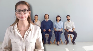 Beyaz gömlekli ve gözlüklü bir kadın, sandalyelerde oturan bir grup insanın önünde durmaktadır. Kadın onlarla konuşuyor gibi görünüyor. Ön planda bir adam bir taburede oturmaktadır. Onun sağında bir başka kişi sandalyede oturmakta ve elinde bir kâğıt tutmaktadır. Diğer tarafta bir adam sandalyede oturmuş, elinde bir tablet tutuyor. Çerçevede bir kolyenin yakın çekimi de görülüyor. Arka plan hafif bulanıkken, sandalyelerde oturan insan grubu odakta. Sağ alt köşede, sandalyede oturan bir kişinin bacakları görülebiliyor.