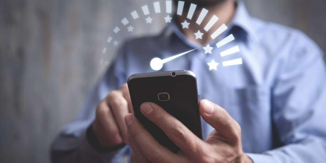 A person is standing holding up a white smartphone in their hands. They have a determined expression on their face, with their mouth slightly open and their eyes looking down at the device. Their index and middle fingers are slightly bent, gripping the device securely. The phone has a thin black border and a smooth glass screen, with the home button in the center. Their arm is straight, with their hand close to their chest. They are wearing a gray t-shirt, blue jeans, and white sneakers. The background is a neutral gray wall.