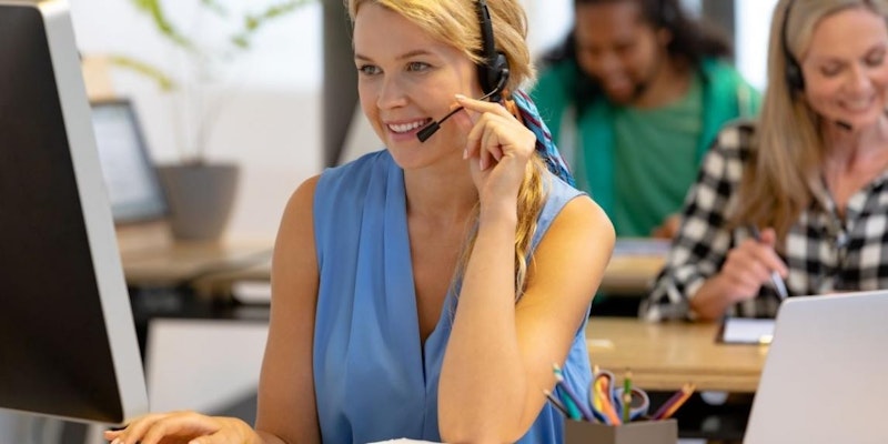 Kulaklık takan bir kadın bilgisayar ekranına bakmaktadır. Açık mavi bir kafa bandı olan siyah bir kulaklık takıyor ve saçları düzgünce arkadan bağlanmış. Yüzünde ciddi ama odaklanmış bir ifade var ve gözleri önündeki ekrana sabitlenmiş. Sağında bir dizüstü bilgisayar var ve arka planda beyaz bir nesne ve fincan görülebiliyor. Kadın yumuşak bir ışıkla aydınlatılmış ve sıcak bir atmosfer yaratılmış. Görüntü keskin ve nettir, genel bir hassasiyet ve doğruluk izlenimi verir.