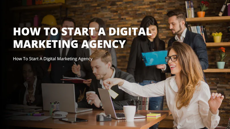 A woman sits at a table with a laptop in front of her and a group of people standing behind her. She appears to be smiling and is wearing a white top with blue jeans. On the table is a laptop, a white cup, and a holder with pens in it. To the right of her is a blue rectangular object with black corners. To the left of her is a potted flower. Additionally, there is a man and woman in the room holding a clipboard and a yellow paper respectively. The woman has a yellow paper in her hand as well.