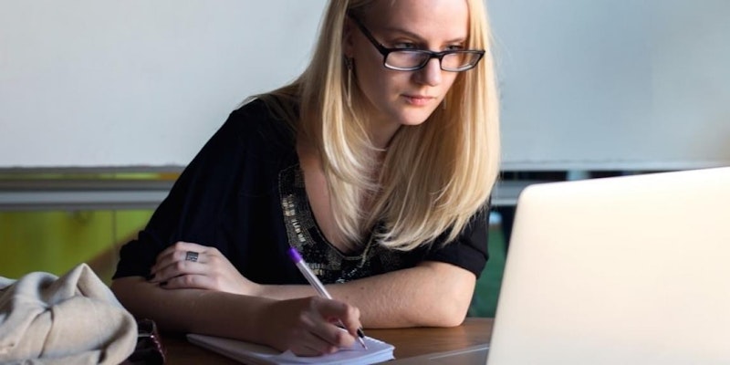 Siyah bir kalem tutan ve beyaz bir deftere yazı yazan bir kadın elinin yakın çekimi. Defter açık ve kağıt hafifçe bulanık. Kalem kağıda doğru tutulmuş ve kadın işine odaklanmıştır. Arka plan odak dışıdır ve arka planda gri ve siyah kasalı bir dizüstü bilgisayar görünmektedir. Görüntünün ön planında beyaz bir yastık var. Kadın kısa kollu beyaz bir gömlek giymiş ve saçlarını yüzünden geriye doğru toplamış.