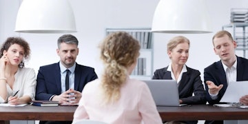 Bir grup insan profesyonel bir ortamda büyük dikdörtgen bir masanın etrafında oturmaktadır. Masanın ortasında bir dizüstü bilgisayar açık ve takım elbiseli bir kadın dikkatle ona bakıyor. Solunda, takım elbiseli ve kravatlı bir adam elinde bir kağıt parçası tutmakta, sağında ise takım elbiseli bir başka adam aynı şeyi yapmaktadır. Masanın arka tarafında, beyaz gömlek giymiş kıvırcık saçlı bir kadın dizüstü bilgisayardan uzağa bakıyor. Onun yanında başka bir kişi elinde bir kağıt parçası tutmaktadır. Masa beyaz bir arka plana sahip ve tüm insanlar ellerindeki işe odaklanmış durumda.