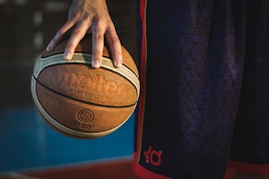 Bir kişi elinde bir basketbol topu tutmaktadır ve top avuçlarının ortasındadır. Basketbol topunun üzerinde yakın çekimde görülebilen beyaz ve kırmızı bir logo vardır. Kişi üzerinde kırmızı bir harf bulunan siyah bir gömlek giymektedir. Görüntünün arka planında mavi bir havlu bulunmaktadır. Kişinin eli odakta ve yakın çekimde bir parmak ucu görülebiliyor. Basketbol topu kameraya doğru tutulmakta ve logoyu göstermektedir.
