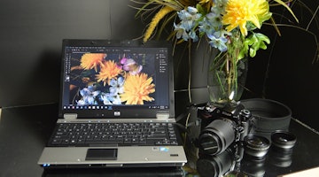 Bej renkli bir duvarın önündeki masanın üzerinde siyah bir dizüstü bilgisayar duruyor. Masanın üzerinde içi çeşitli parlak sarı ve mor çiçeklerle dolu beyaz bir vazo var. Dizüstü bilgisayarın ekranı açık ve klavyesi görülebiliyor. Dizüstü bilgisayarın üzerine büyük lensli bir kamera yerleştirilmiş. Kameranın siyah bir gövdesi ve parlak bir merceği vardır. Dizüstü bilgisayarın köşesinde, yaprakları dışa doğru yayılan sarı bir çiçeğin yakın çekimi yer alıyor. Bir klavyenin ve bir kamera lensinin yakın çekimi de görülebiliyor. Dizüstü bilgisayar, çiçekler ve kamera, dijital ve doğal unsurların ilginç bir yan yana gelişini oluşturuyor.