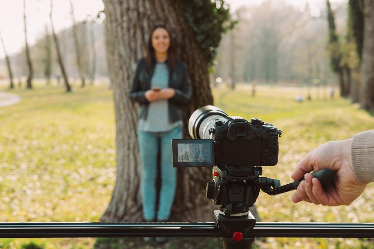 Gri gömlek giyen bir kişi siyah bir dijital fotoğraf makinesini bir kadının yüzüne doğru tutuyor. Kadının uzun, siyah saçları var ve mavi bir bluz giyiyor. Ayrıca sol elinde beyaz bir telefon tutmaktadır. Arkalarında tripod üzerinde bir kamera görülüyor ve ön planda bir kişinin bacağının bulanık bir görüntüsü var. Arka planda, ağaçların ve birkaç binanın bulunduğu bir parkın bulanık bir görüntüsü var.