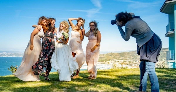 Hepsi de güzel beyaz elbiseler giymiş bir grup kadın fotoğraf için poz veriyor. Kadınlar birbirlerine sımsıkı sarılmış, ışıl ışıl gülümsüyor ve kameraya doğru bakıyorlar. Yan tarafta duran bir adam grubun fotoğrafını çekerek bu mutlu anı yakalıyor. Arkalarında, ağaçların ve dağların uzandığı bir manzara uzanıyor. Özellikle bir kadın önde duruyor, elbisesi rüzgarda dalgalanıyor ve yakın çekimde düzgün bacaklarını ortaya çıkarıyor. Fotoğrafın odak noktası olan kadın grubu, birlikte geçirilen özel bir günün kalıcı bir hatırlatıcısı.