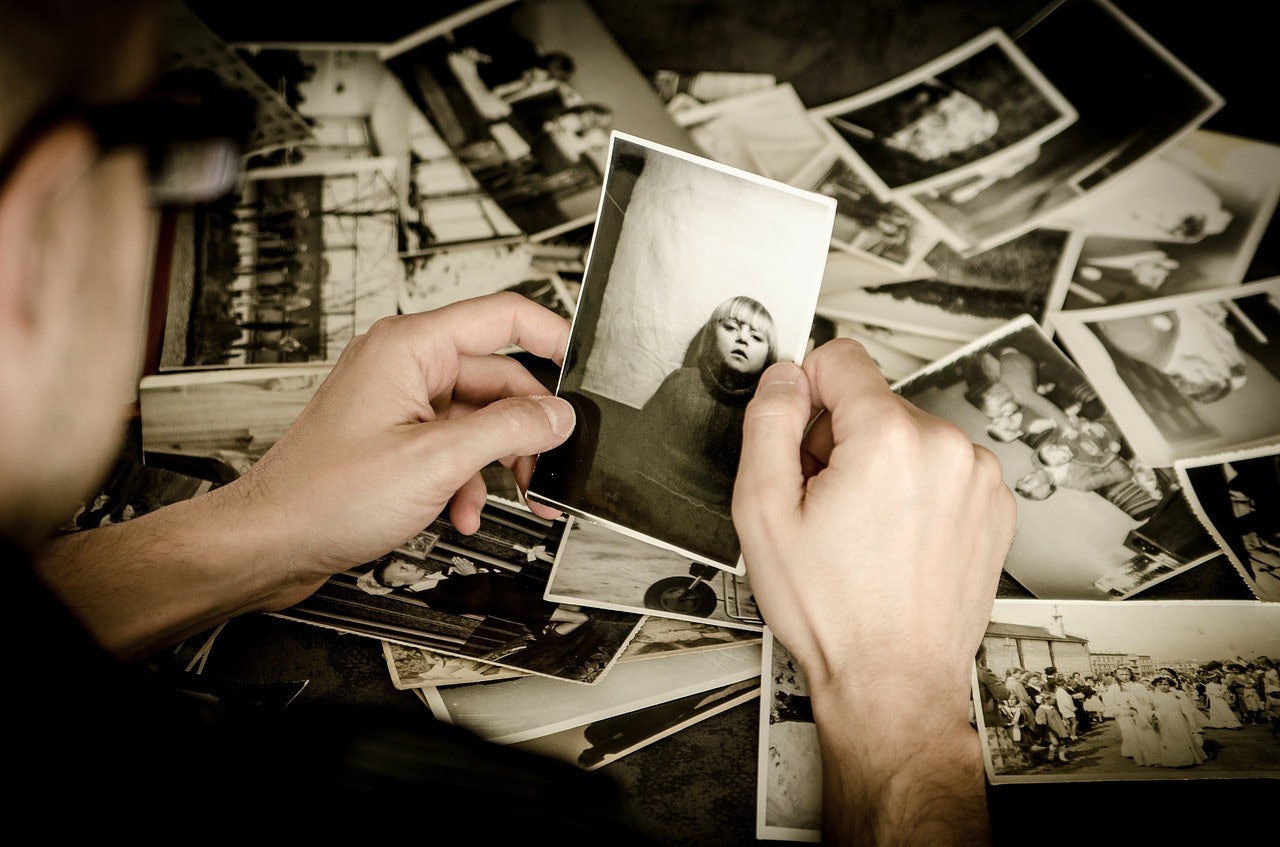 Beyaz elbise giyen bir kişi, kameranın çekebilmesi için gülümseyen bir çocuğun fotoğrafını tutuyor. Fotoğraftaki çocuk mavi bir tişört giymektedir ve yüzünde parlak bir gülümseme vardır. Fotoğrafı tutan kişi diğer elini kalçasına koymuş ve doğrudan kameraya bakmaktadır. Fotoğraf yakın plandan çekilmiş, odak noktası kişi ve tuttuğu fotoğraf. Arka plan bulanıktır ve arka planda bir yığın başka fotoğraf görülebilir. Ayrıca, görüntünün sağ alt köşesinde bulanık bir tekerlek görülebiliyor.