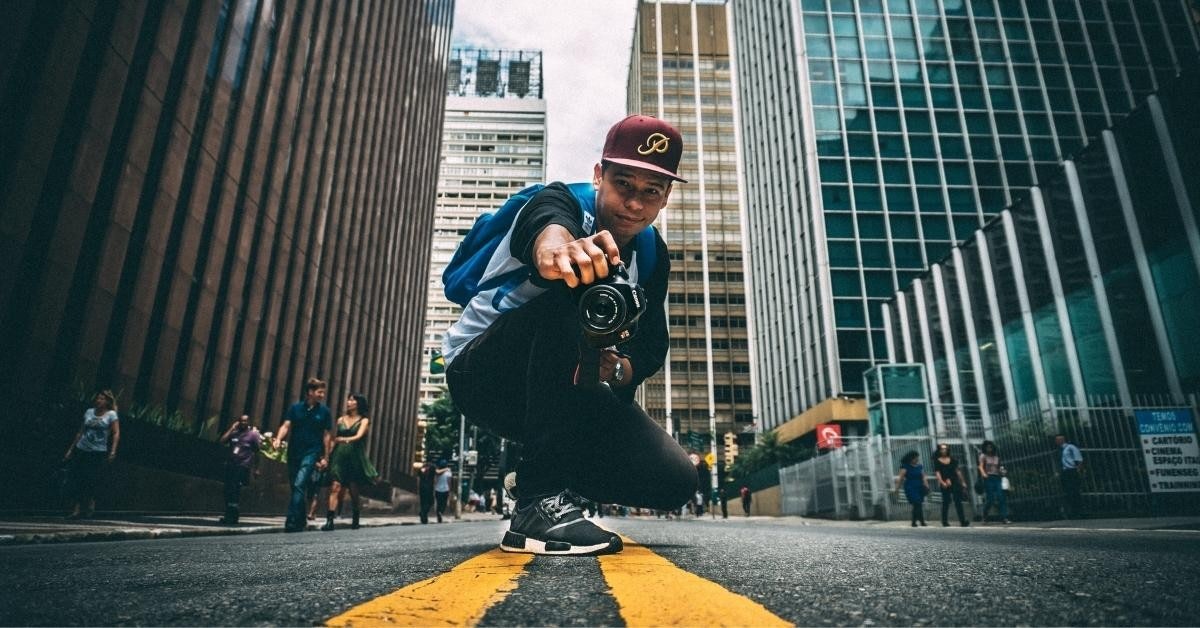 A man is squatting on the street with a camera in his hands. He is wearing a hat and has a lens of a camera close to his face. His feet are on a road and he is walking in a blurry image. There is a woman in a green dress in the background, and a group of people walking in front of a tall building. The man is wearing shoes and has a close-up of a shoe on the ground. The street is empty and the sun is shining. The sky is clear and the air is fresh. The picture is taken from a close distance, allowing the viewer to see the details of the man and his camera. The colors are vibrant and the lighting is perfect. The scene is peaceful and serene.