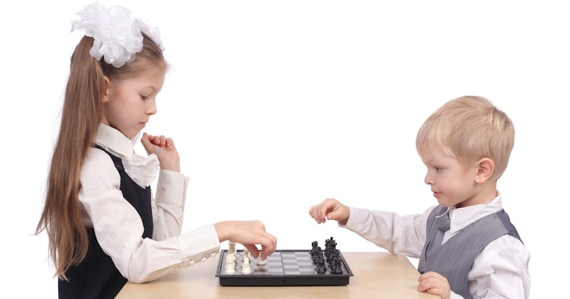 Bir tahta üzerinde satranç oynayan iki kızın yakın çekimi. Kızlardan birinin uzun kahverengi saçları at kuyruğu şeklinde ve beyaz bir çiçek tokasıyla tutturulmuş. Diğer kızın saçları topuz yapılmış. Her ikisinin de saçlarında fiyonk vardır. Kızlardan biri beyaz bir yelek, diğeri ise gri bir tişört giyiyor. Ön planda bir oğlan çocuğu yüzünde konsantre bir ifadeyle satranç oynuyor. Görüntüde kolu görünüyor. Tahtanın bir tarafında siyah, diğer tarafında beyaz taşlar var.