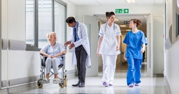 Bu görüntüde bir hastanede yürüyen bir grup insan tasvir edilmiştir. Fotoğrafın odağında mavi önlüklü bir kadın var ve fotoğrafın ortasında yürüyor. Solunda tekerlekli sandalyedeki bir kadın, sağında ise beyaz önlüklü ve beyaz pantolonlu bir kadın görülüyor. Daha sağda, beyaz önlüklü bir adam bir hastayla konuşurken görülüyor. Mavi önlüklü kadının yakın çekimi yüzünü ortaya çıkarıyor ve bir kişinin bacağının yakın çekimi de görülebiliyor. Ayrıca arka planda sandalyede oturan bir kişi görülüyor. Bu fotoğraf, modern dünyada sağlık hizmetlerinin önemini vurgulayarak yoğun bir hastanenin koşuşturmasını yansıtıyor.
