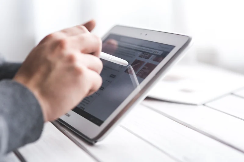 Resimde tablet bilgisayar kullanan bir kişi gösterilmektedir. Cihazı sağ elinde tutmakta, diğer elinde ise bir kalem bulunmaktadır. Tablet beyaz renkte, ekran gri bir arka plana sahip ve üzerinde bir nesnenin yakın çekimi var. Nesne beyaz gibi görünmektedir ancak bulanıktır. Kişi tabletle etkileşime geçmek için kalemi kullanmaktadır. Sahnenin geneli iyi aydınlatılmış ve kişi ile cihaz arasında iyi bir kontrast var. Arkalarında karanlık bir alan var ve görüntü için güzel bir fon oluşturuyor.