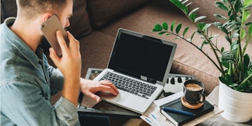 Bir adam masasında oturmuş, elindeki telefona odaklanmış. Önünde açık duran dizüstü bilgisayar siyah renktedir ve yan tarafında kahverengi bir sıvı dolu bir kupa durmaktadır. Sol tarafta yeşil yapraklı beyaz bir vazo, sağ tarafta ise üzerinde kahve fincanı olan bir kitap duruyor. Gri bir kapüşonlu sweatshirt ve altında siyah bir tişört giyiyor. Kısa, koyu renk saçları var ve dikkatle telefona bakıyor. Ciddi bir konuşma yapıyor gibi görünüyor.