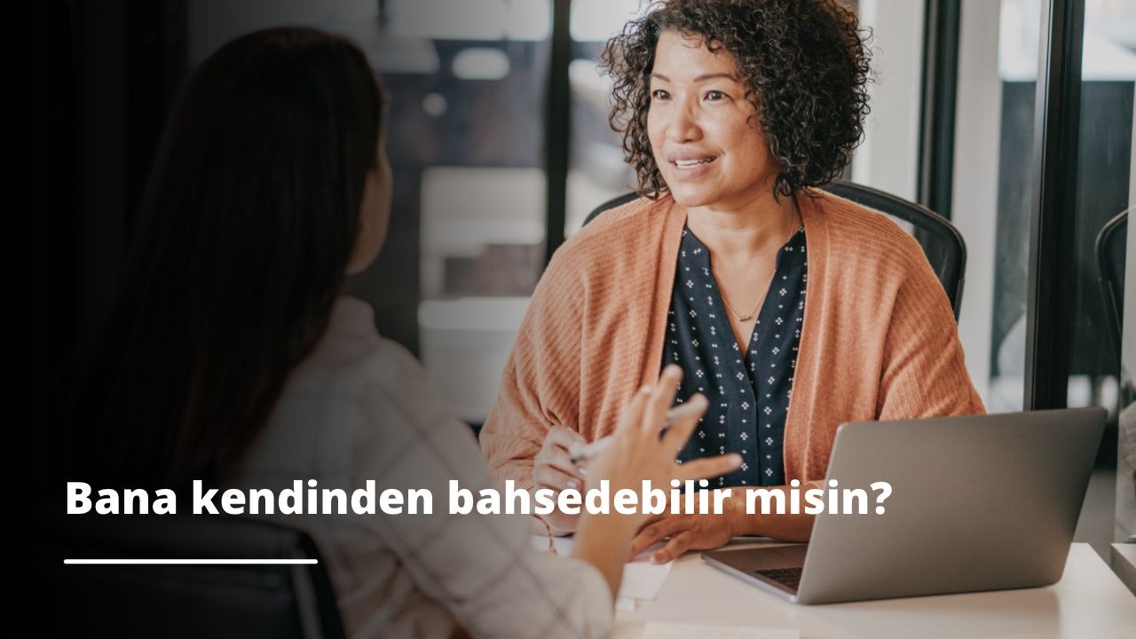 Genç bir kadın önünde dizüstü bilgisayarıyla bir masada oturuyor. Gözleri ekrana odaklanmış ve bir şeyler yazıyor. Kolyesi takılı ve kahverengi bir kazak giyiyor. Yüzü yakın plandadır ve konsantrasyon ifadesini göstermektedir. Uzun kahverengi saçları dizüstü bilgisayar tarafından çerçevelenmiştir. Elleri klavyenin üzerinde ve bir şeyler yazıyor gibi görünüyor. Rahat bir pozisyonda ve yaptığı işten keyif alıyor gibi görünüyor. Derin düşüncelere dalmış, önemli bir şey üzerinde çalışıyor gibi görünüyor.